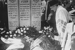 Cantor Pinchos Rabinowitz, clad in a large prayer shawl, offers prayers in front of a memorial to the victims of the Holocaust adorned with wreaths of flowers.