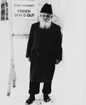 An elderly Jewish man stands next to a sign in the Hasenecke DP camp that reads, "Review Concert -- It's Good to be Happy."