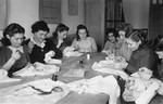 A group of women sit around a table and hand-sew garments in an ORT (Organization for Rehabilitation through Training) workshop in the Landsberg displaced persons' camp.