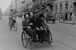 Three Jewish men ride in a rickshaw along a street in the Warsaw ghetto.