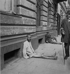 A destitute man wearing nothing but a shirt sits on the pavement near the street entrance to the Pinkiert funeral home in the Warsaw ghetto.