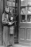 A destitute Jewish woman poses in the doorway of a store in the Warsaw ghetto holding a metal container.