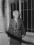 An elderly female survivor poses in front of a barred window at the Hadamar Institute, where she was imprisoned for writing anti-Nazi articles.