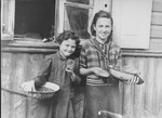 Two young girls stand outside a house in the Kovno ghetto holding bread and bowls of food.