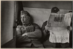 Two German men sit in a train car; one is reading a newspaper.
