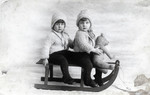 Studio portrait of twins Regina and Ruth Anders sitting on a toy sled with a stuffed bear.