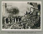 Men and women stand behind piles of rubble in a bombed out city quarter.