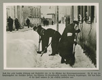 Three men, one with an armband, clear snow in the street of an unidentified ghetto.