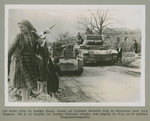 German tanks roll down on a dirt road. while men, women, and children stand offside.