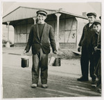 A young man carries lunch pails in each hand outside barrack #5 in the Ziegenhain displaced persons camp.