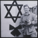 Portrait of photographer Phil Drell standing with his camera in front of a large Jewish star erected for the Dachau camp memorial.