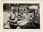 Two young children sit at an outdoor table by a fence.