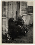 Two destitute women sit against a fence in the Lodz ghetto wrapped in blankets.
