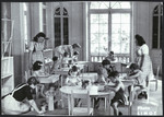 Young children, supervised by female staff, sit and play at tables in a classroom of a postwar OSE children's home.