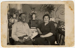 A Jewish couple sits around a small table covered with an embroidered tablecloth in their home.