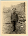 A child survivor in a uniform stands smiling amid the rubble of Nordhausen concentration camp.