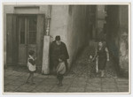 A religious Jew leans against a building while a woman and children walk and play nearby.