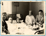 Two young men lie in a hospital bed in a hospital surrounded by family and friends.