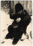 A destitute young boy in tattered clothes is eating his soup seated in the snow in the Lodz ghetto.
