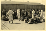 A group of men line up outside a building; they are being attended by five men dressed in white overalls.
