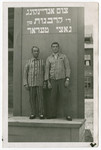 Two Jewish displaced persons wearing their concentration camp uniform pose in front of a memorial to the victims of the Holocaust.