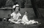 Two Jewish sisters sit on the grass next to a baby stroller.