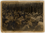 A large group of men is seating on the ground with their hands on their heads while being forced to watch the execution of Moshe Kagan and Wolf Kieper.