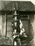 Five young male students posie on the rungs of a ladder outside of a building that looks like a barn or shed.