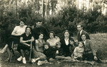 A group of men. women and children sit outside by a wooded area.