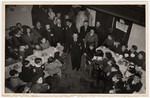 Children in a postwar children's home gather around long tables set with fruit and decorated with flowers.