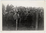 Survivors wearing blankets for warmth crowd together behind a barbed wire fence shortly after liberation.