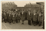 Children in uniform stand at attention in the Aschaffenburg displaced persons' camp while a band plays in the background.