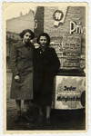 Rochen (Rosi) and Tosca stand in front of a pillar covered with posters and advertisements.