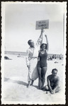 A man and woman hold up a sign reading while vacationing on a beach.