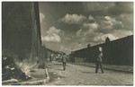 Survivors walk past the barracks of the Buchenwald concentration camp while a small fire burns on the side.