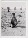 A small boy [probably Spanish] crouches down, as he plays in the dirt in front of the barracks of the Rivesaltes internment camp.
