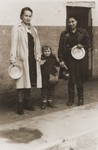 Two women and a child pose in front of a soup kitchen in the Cremona displaced persons camp holding metal bowls.