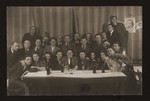 Group portrait of Jewish DPs in the Heidenheim displaced persons camp gathered around a table next to a portrait of Revisionist Zionist leader, Vladimir Jabotinsky.