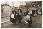 Elderly women carrying young children and bundles of personal belongings trudge along a street in the Lodz ghetto toward the assembly point for deportations to Chelmno.