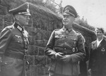 Two high-ranking German military officers converse in front of a stone wall.