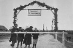 Three men and a woman stand behind the guard rail at the entrance to the Maly Trostinets concentration camp.