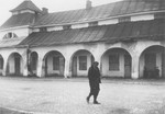 A destitute Jewish man in a torn jacket with an armband walks past a large building in an unidentified town in Poland.
