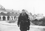 A destitute Jewish man in a torn jacket with an armband stands outside a large building in an unidentified town in Poland.
