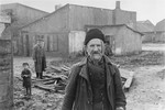 An elderly Jew stands in an open area between buildings in the Kovno ghetto, in front of a pile of wood.