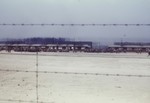 View through the barbed wire fence of survivors gathered outside a row of barracks in the newly liberated Buchenwald concentration camp.