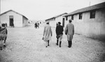 Three [visitors] walk along a street between rows of barracks in the Rivesaltes internment camp in France.