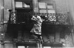 A woman hangs from a balcony, preparing to drop to the street and the waiting SS.
