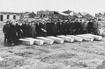 Mourners stand behind a row of coffins at an unidentified  burial site

The location of the photograph has been tentatively identified as the Jewish Cemetery in Kielce, Poland, with buildings on Dzika Street visible in the background.
