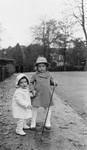 Two young Jewish children stand on the sidewalk dressed in hats and coats.