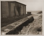A partially filled mass grave lies in the foreground as American soldiers walk around a barn outside of Gardelegen in which over 1,000 prisoners were burned alive by the SS.